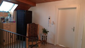 Une pièce avec un plafond en bois, une porte blanche, une chaise en osier, une petite table avec une plante et un bureau sur lequel se trouvent divers objets.