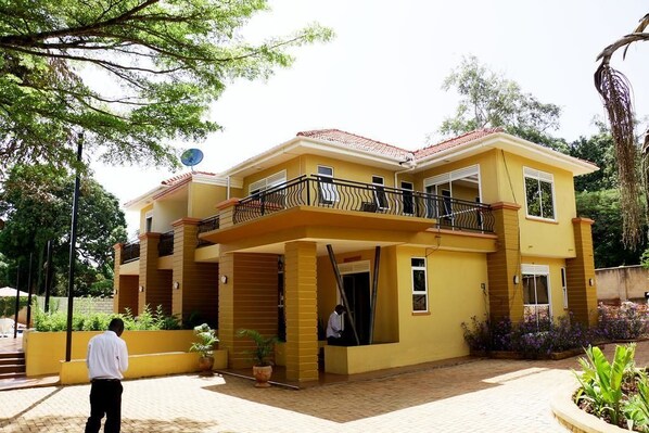 A two-story yellow building with a balcony, a satellite dish, and a paved entrance area with potted plants. - The Mansion Hotel (Jinja)