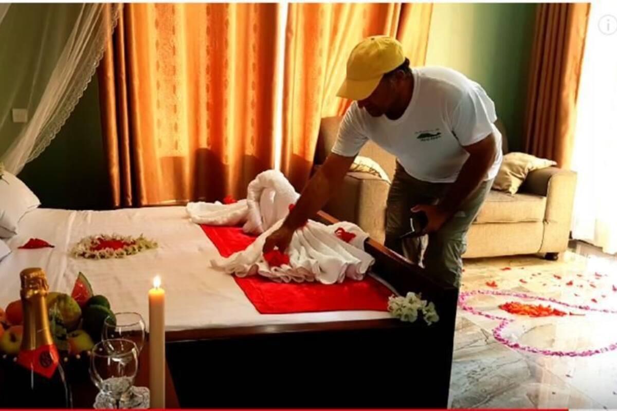 Un lit bien fait, avec une couverture rouge, des oreillers blancs et un bouquet de fleurs. On y trouve également des bougies, un panier de fruits et une bouteille de champagne sur la table de chevet.