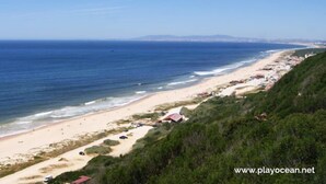 A coastal beach with a sandy shore, a few umbrellas, and a few buildings along the coastline. - Backpackers Hostel Fonte da Telha (Almada)