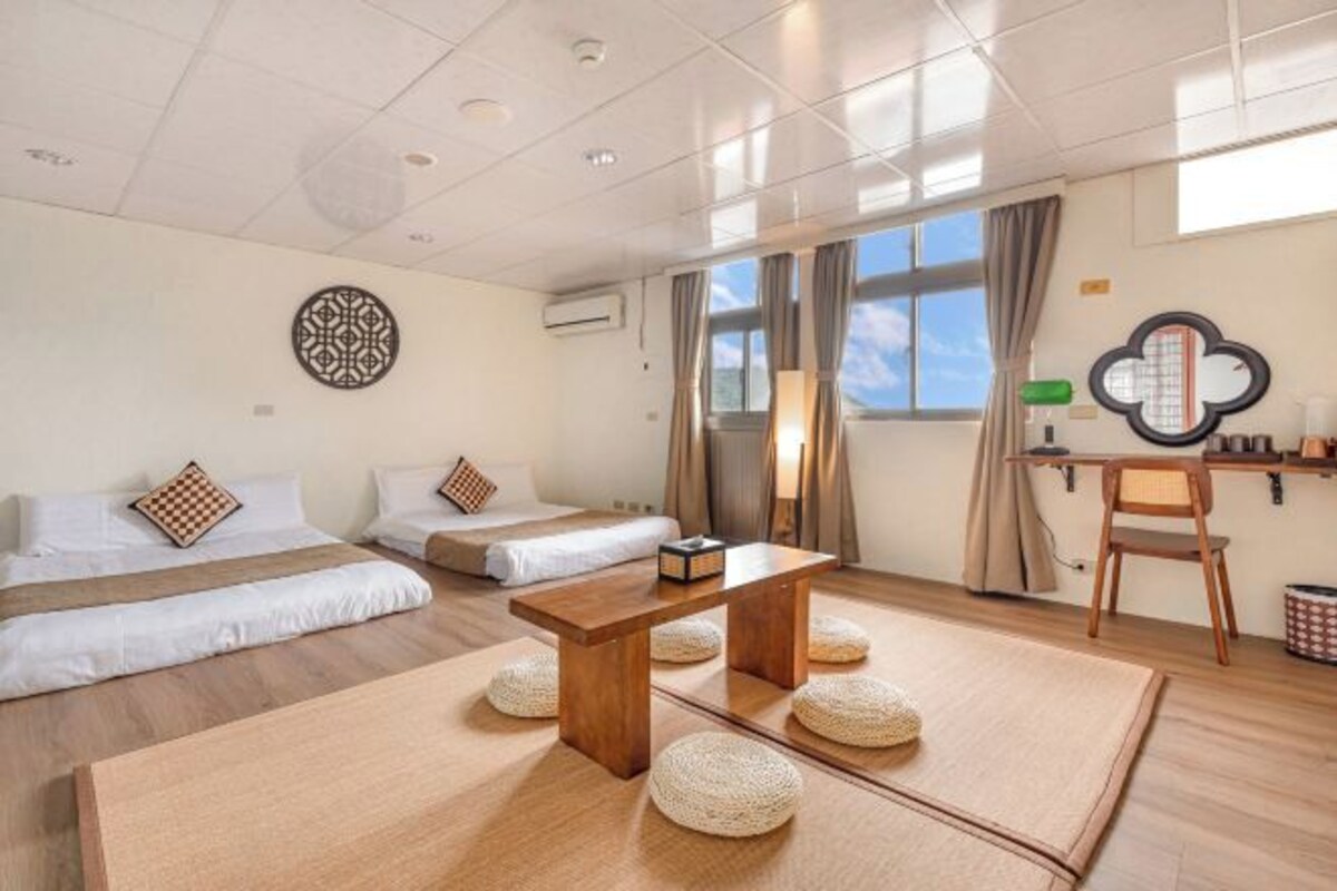 A modern hotel room with two beds, a wooden table, and a large window with curtains.