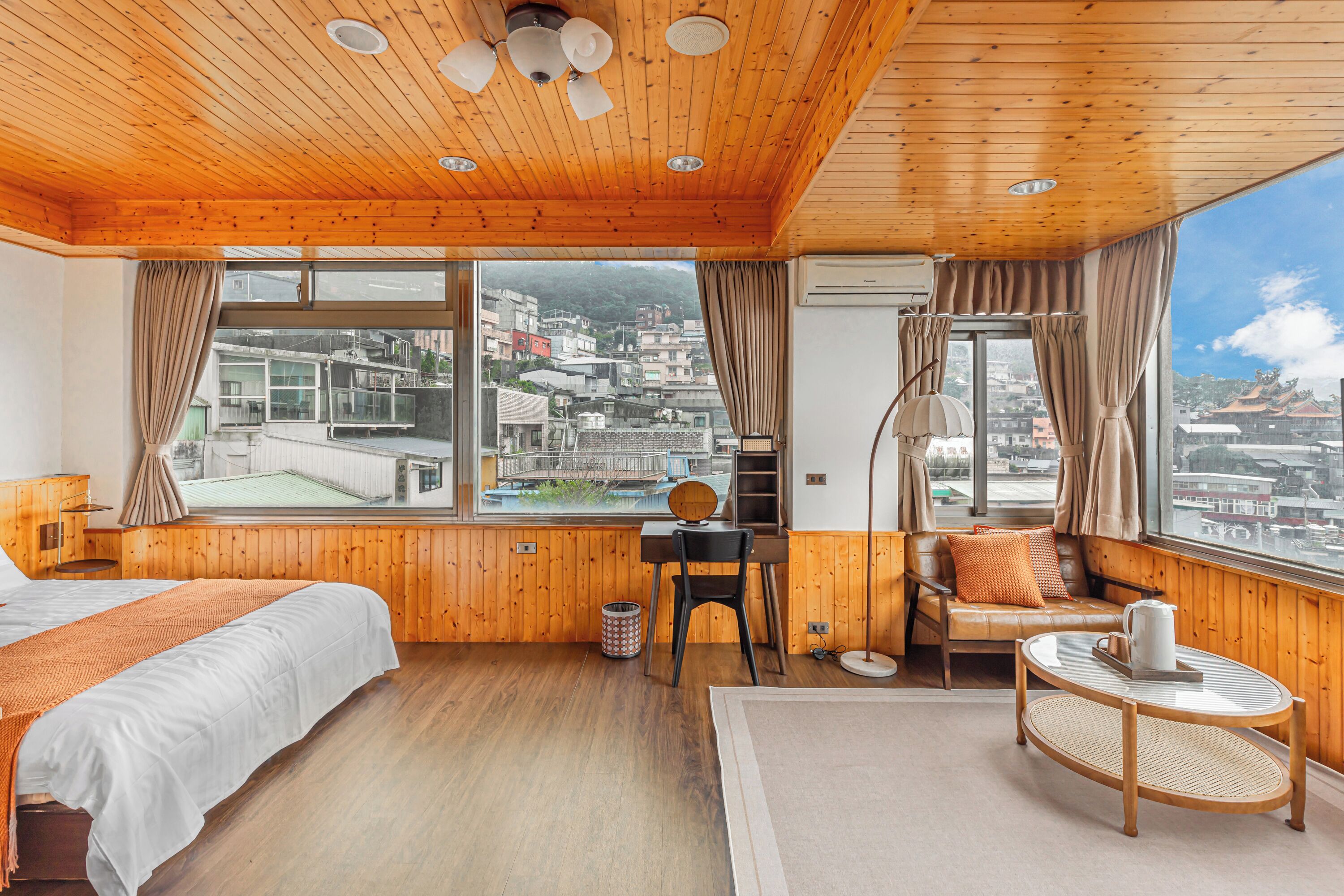 Habitación de hotel con paredes y techo de madera, una ventana grande con vistas a la ciudad, una cama, un escritorio con una silla, un sofá y una mesita redonda.