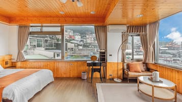 Habitación de hotel con paredes y techo de madera, una ventana grande con vistas a la ciudad, una cama, un escritorio con una silla, un sofá y una mesita redonda.