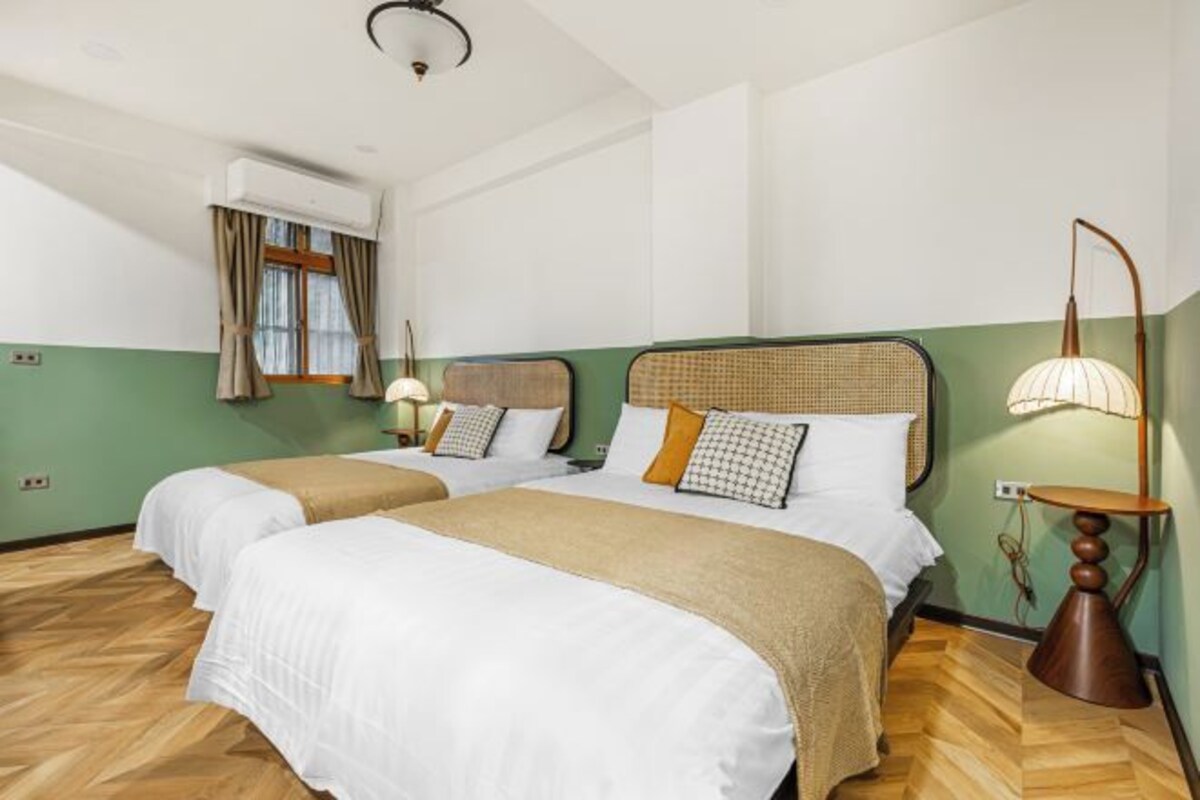 A hotel room with two beds, a window with curtains, a standing lamp, and a wall-mounted air conditioner.