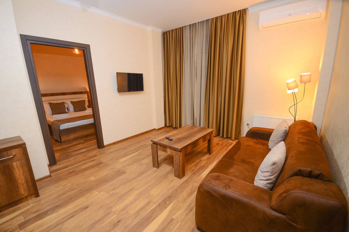 A room with a wooden floor, a brown sofa, a wooden coffee table, a TV mounted on the wall, and a bed in the background.