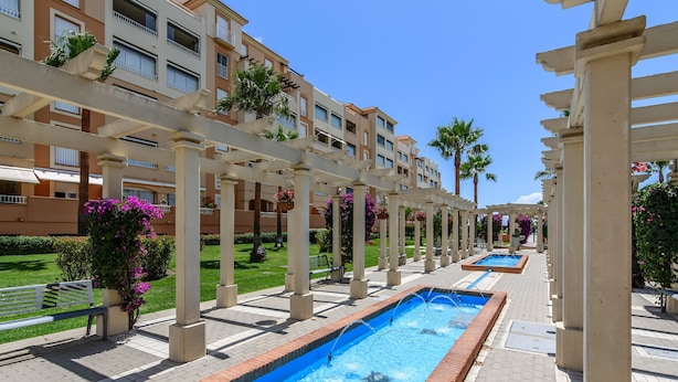 A pool area with a pergola structure, a water feature, and a row of residential buildings in the background. - Las Arenas - Haraganes Beach HLHA181P193 (Isla Canela)