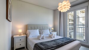 A modern bedroom with a tufted headboard, a large window with a view of the building, and a hanging light fixture. - Las Arenas - Haraganes Beach HLHA181P193 (Isla Canela)