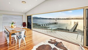 A spacious room with a large wooden dining table, white chairs, a glass door leading to a balcony with a view of a lake and pier. - Peaceful Waterfront house Great View Private Jetty (Gosford)