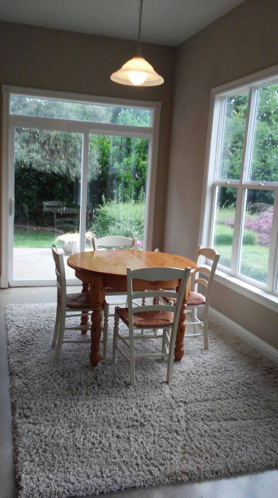 A dining area with a wooden table and chairs, a hanging light fixture, and large windows with a view of greenery.