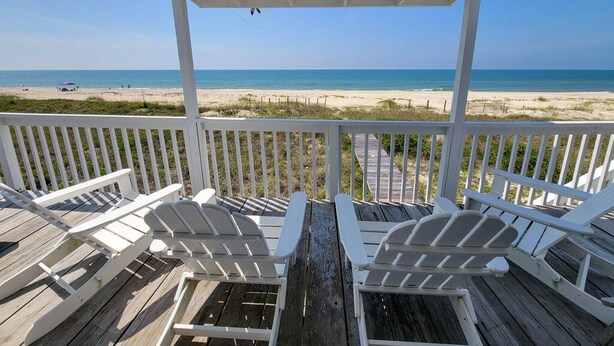 Una terraza de madera con sillas Adirondack blancas que dan a la playa.
