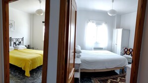 Ein Hotelzimmer mit zwei Betten, eines mit einer gelben Tagesdecke und das andere mit einer weißen. Es gibt ein Fenster mit weißen Vorhängen, einen weißen Kleiderschrank und einen gemusterten Teppich auf dem Boden.
