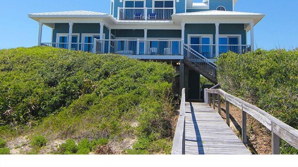 Une grande maison à plusieurs étages, située en bord de mer, avec une terrasse en bois donnant sur un balcon.