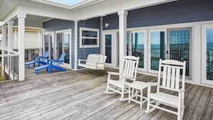 Une terrasse en bois agrémentée de chaises Adirondack blanches et bleues, d’une balançoire et d’une porte vitrée donnant sur un balcon avec vue sur l’océan.