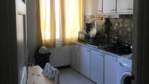Una cocina compacta con armarios blancos, lavadora, una mesa con silla y una ventana con cortinas.
