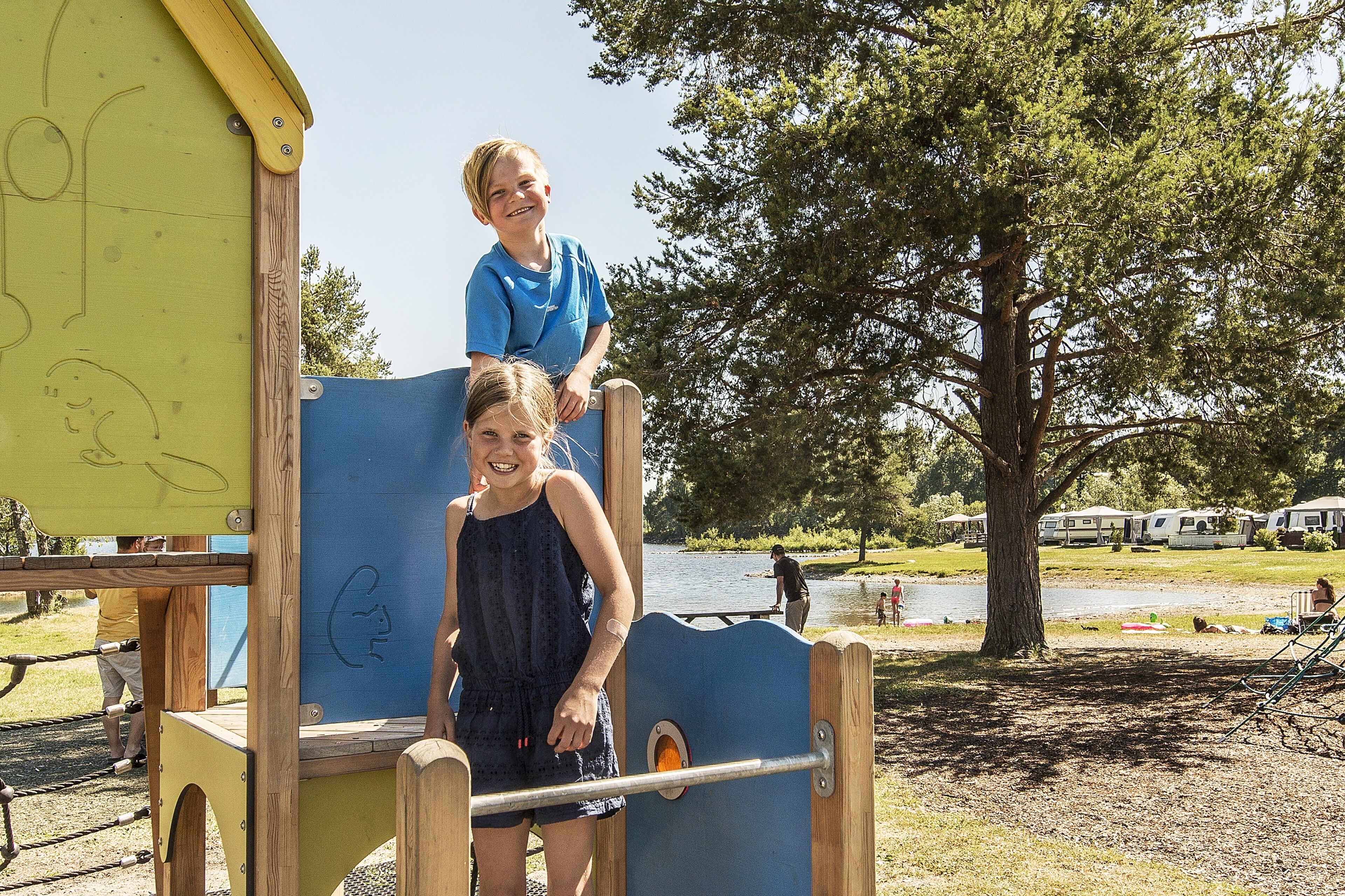 A playground with a wooden climbing structure and a blue slide, set against a backdrop of a lake and RVs.