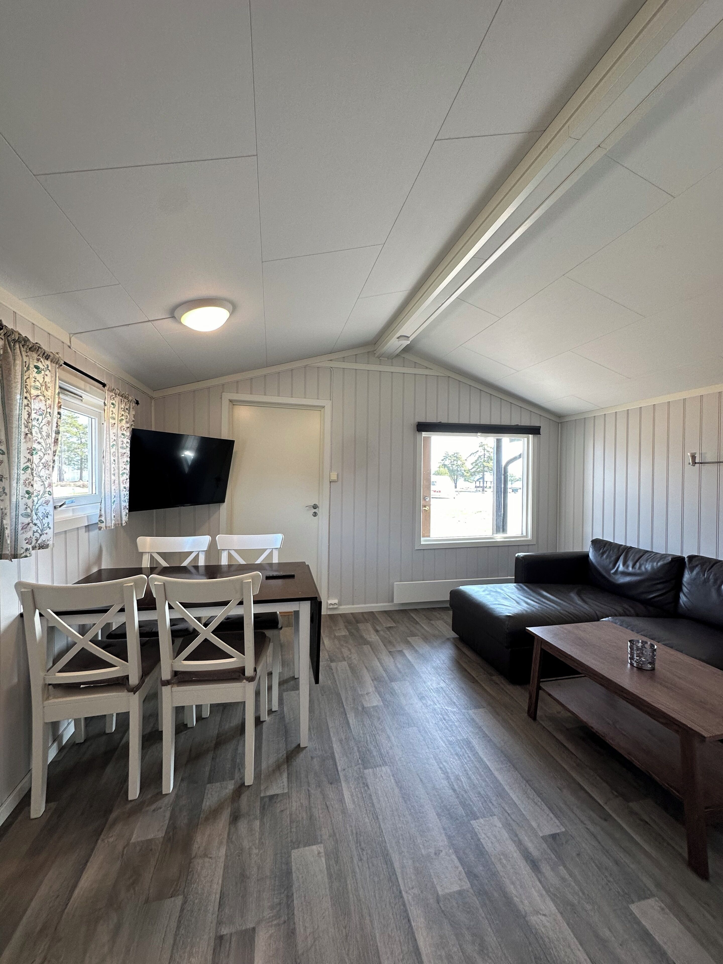Una habitación con una mesa de comedor y sillas, un sofá, un televisor montado en la pared y una ventana con cortinas.