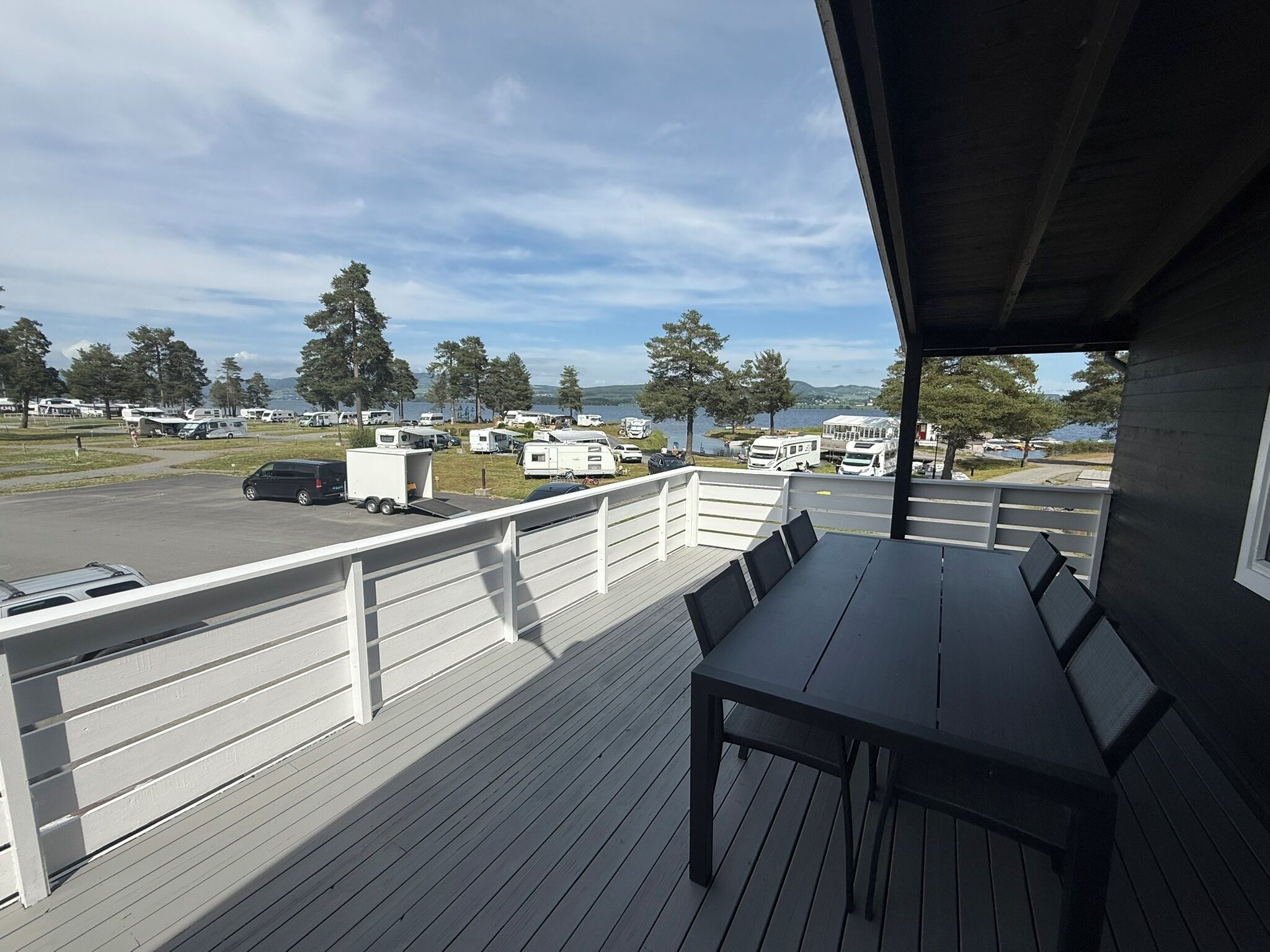 Een overdekt terras buiten met een tafel en stoelen, met uitzicht op een parkeerplaats met campers en een meer.