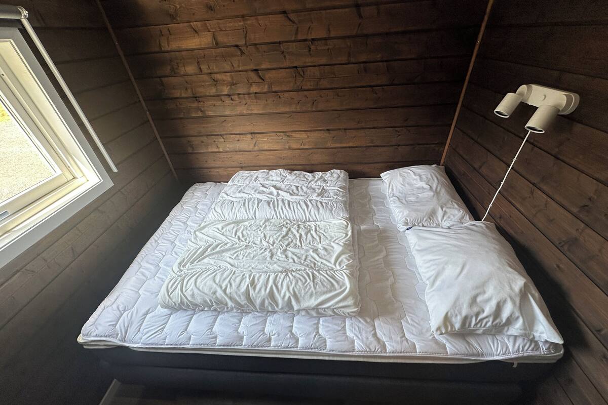 Un lit simple avec un matelas et des oreillers blancs, une tête de lit en bois et une fenêtre sur le côté gauche.