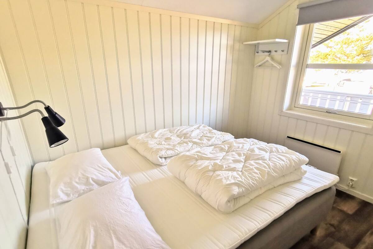 Un lit bien fait, avec du linge de lit blanc, dans une pièce dont les murs sont recouverts de lambris verticaux et qui possède une fenêtre donnant sur l’extérieur.
