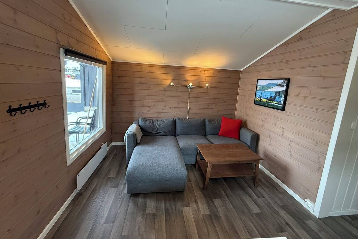 Une pièce chaleureuse avec un canapé d’angle gris, une table basse en bois et une grande fenêtre donnant sur l’extérieur.
