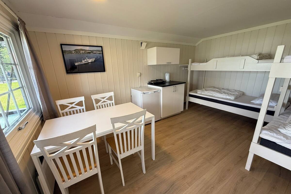 Une pièce compacte comprenant un lit superposé, une petite kitchenette, une table à manger avec des chaises et une fenêtre avec des rideaux.