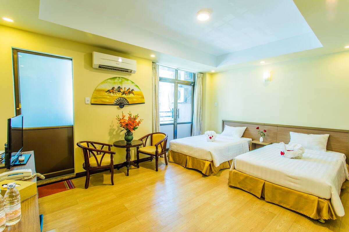 A hotel room with two beds, a desk with a chair, a small table with a vase of flowers, and a painting on the wall.