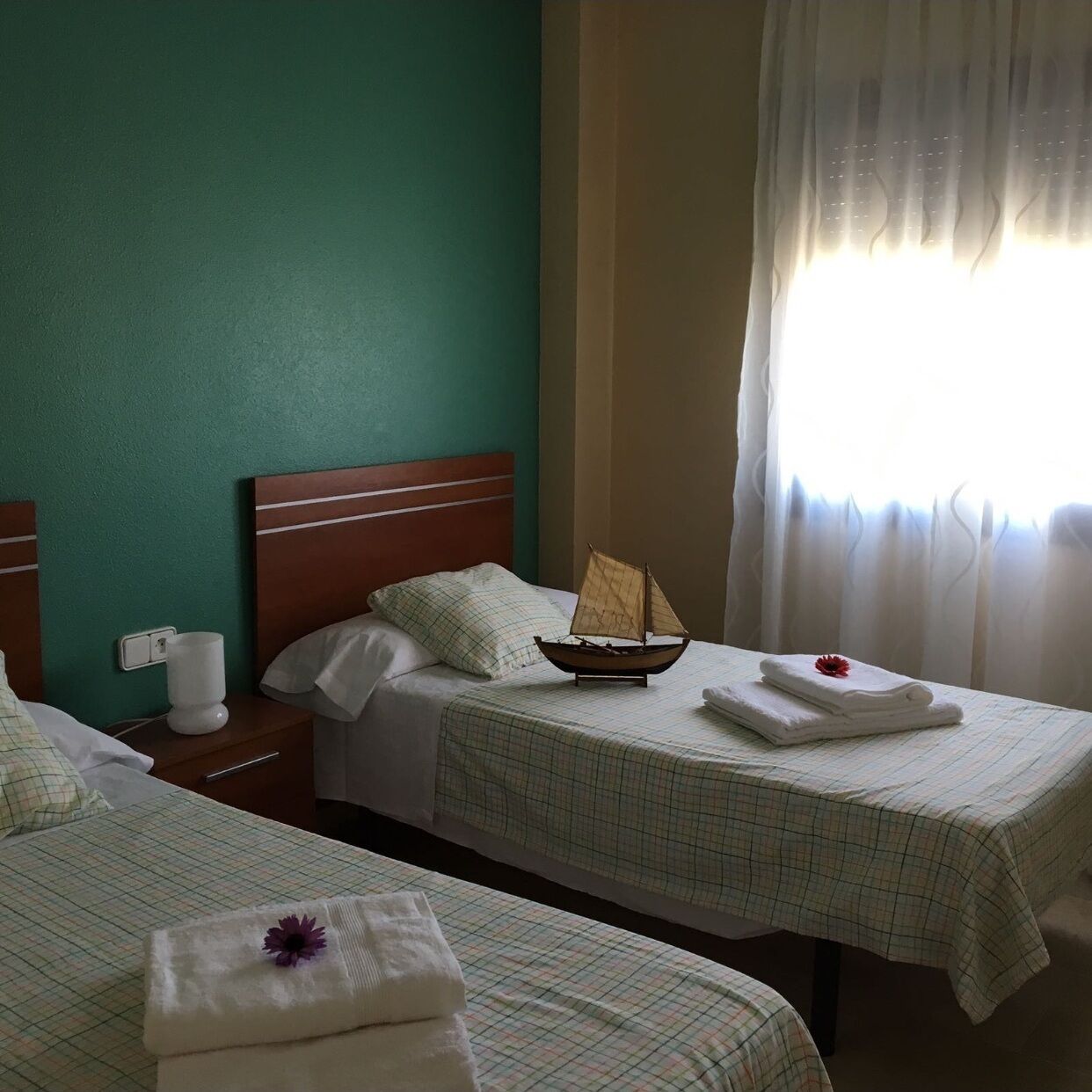 A hotel room with two beds, a wooden headboard, a small boat model, and a window with sheer curtains.
