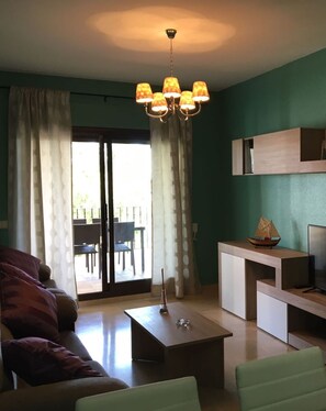 A living room with a sofa, a coffee table, a TV stand, and a chandelier. There is a sliding glass door leading to an outdoor area.