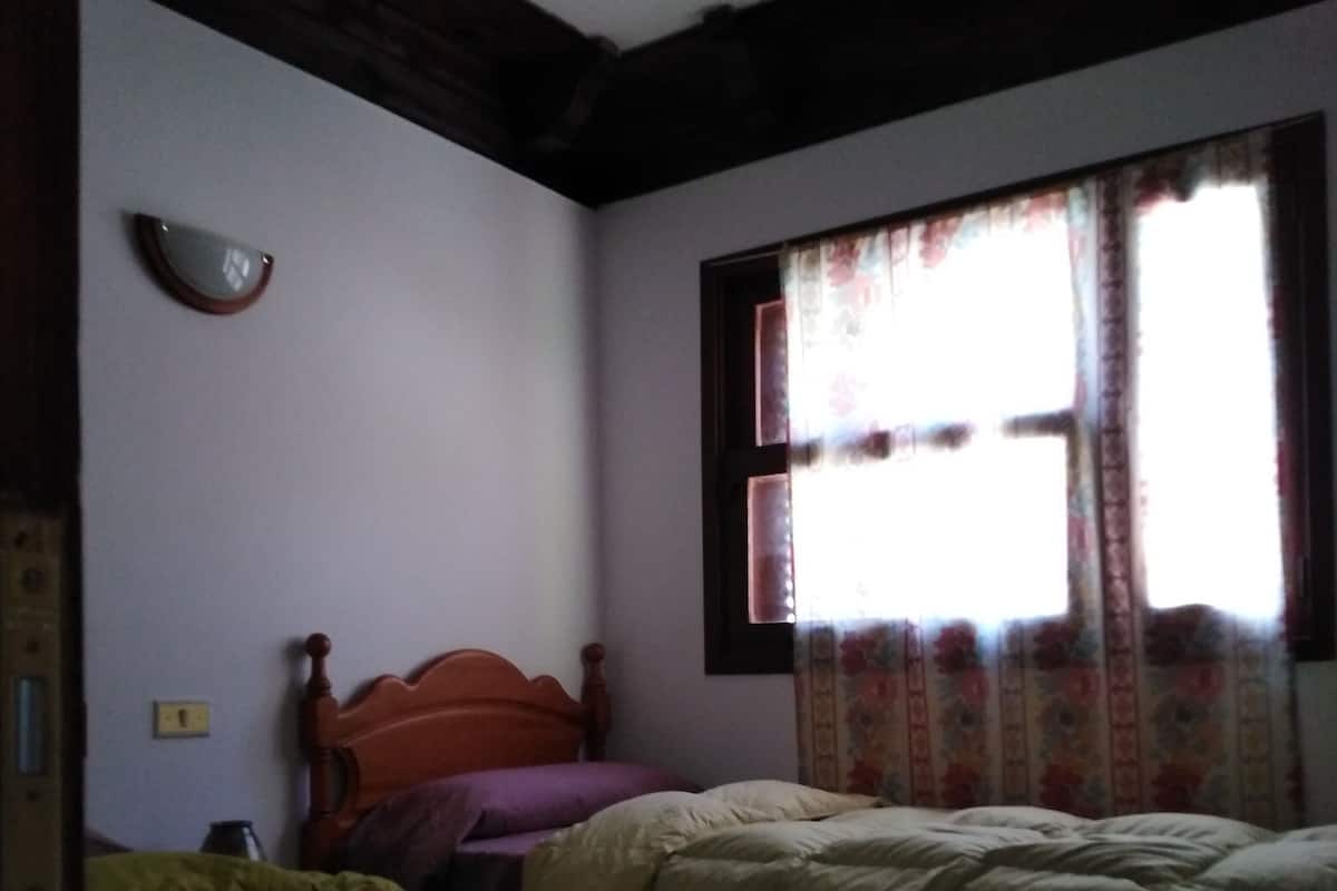 Un dormitorio con dos camas, cabecero de madera, ventana con cortinas florales y una lámpara fijada a la pared.