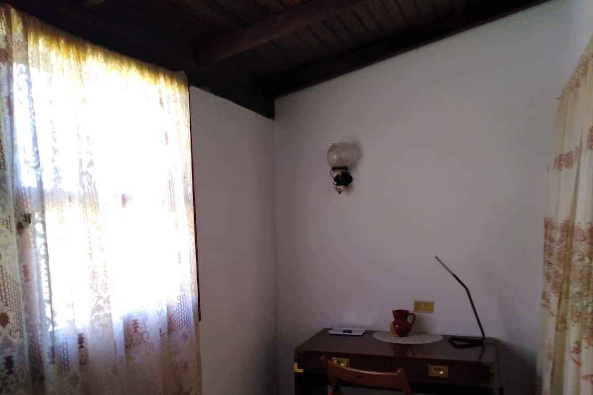 Una habitación pequeña y tenuemente iluminada con una cama individual, un escritorio de madera y una silla. Hay una lámpara fijada a la pared sobre el escritorio.