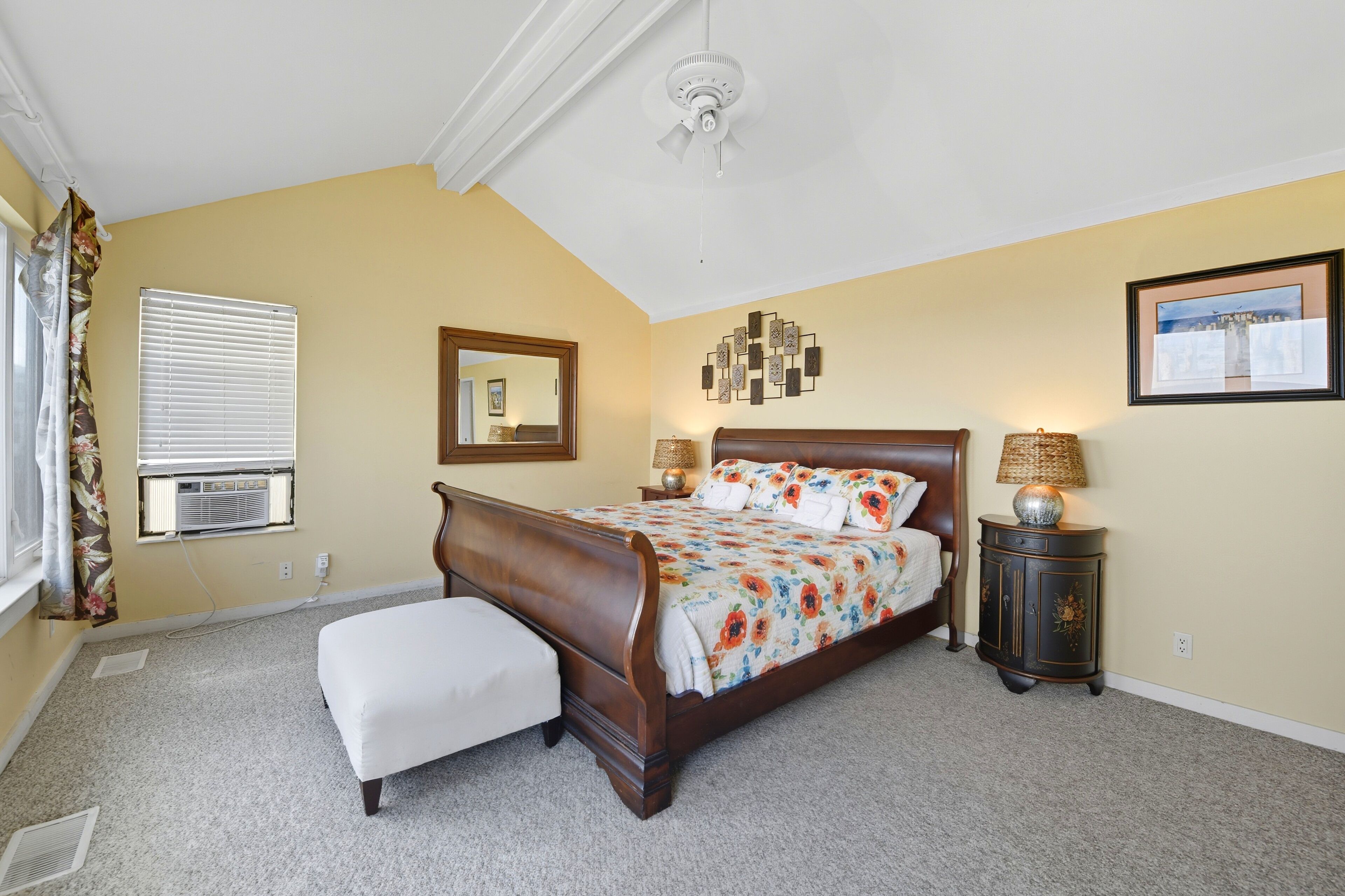 A bedroom with a bed, an ottoman, a nightstand, a lamp, a ceiling fan, a window with blinds, and a mirror.
