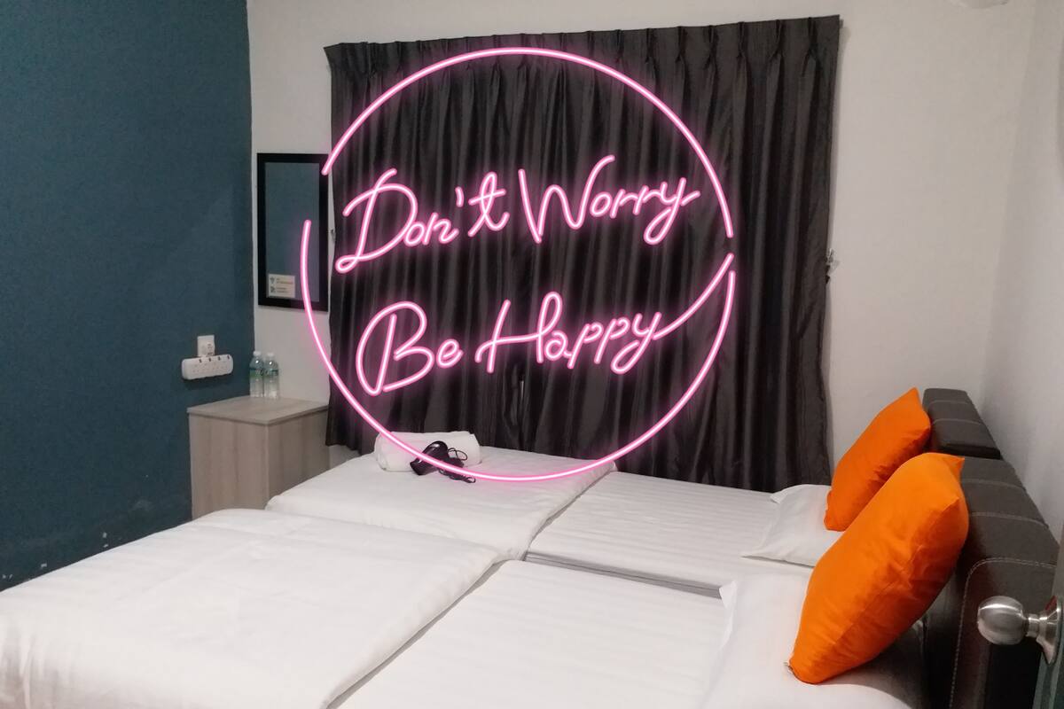 Un lit bien fait, avec des draps blancs et deux oreillers orange, un rideau sombre et un panneau lumineux néon sur lequel on peut lire : « Ne vous inquiétez pas, soyez heureux ».