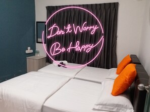 A neatly made bed with white linens and two orange pillows, a dark curtain, and a neon sign that reads "Don't Worry Be Happy".