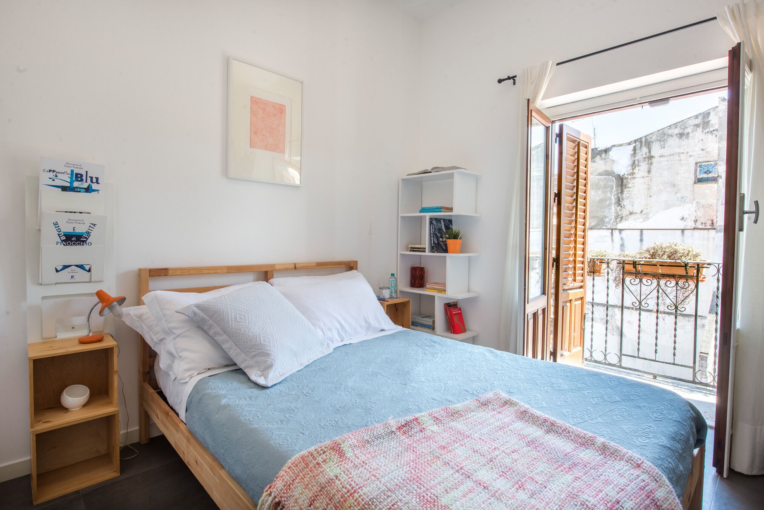 Une chambre à coucher comprenant un lit, une table de chevet, une étagère avec des livres et une plante, et donnant sur le balcon extérieur.