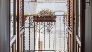Un balcon ouvert avec une grille en fer forgé, une vue sur un immeuble doté d’une fenêtre bleue et un mur en béton.
