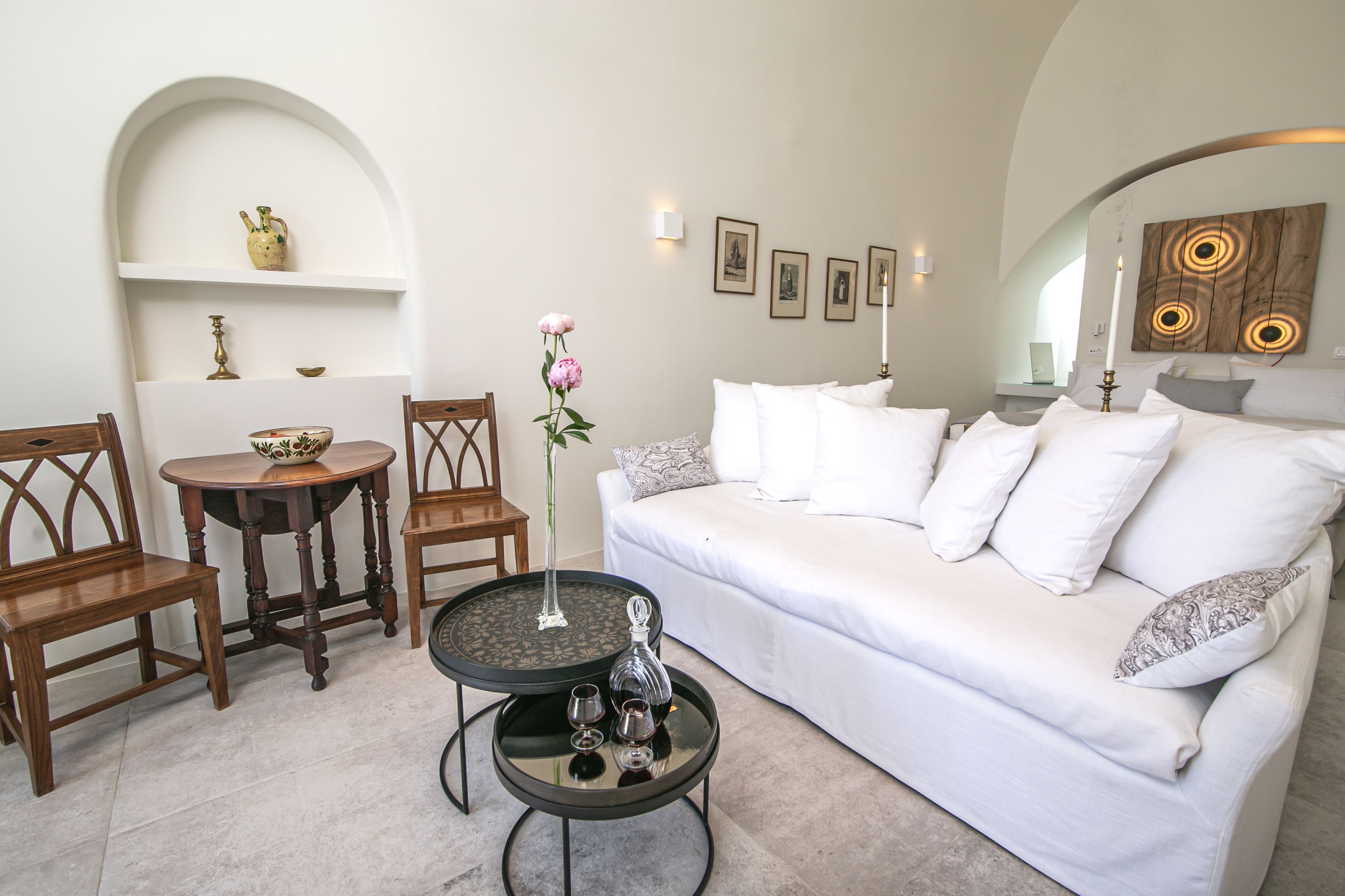 A living room with a white sofa, two wooden chairs, a small round table with a vase and glasses, and a wall with framed pictures and a wooden art piece.