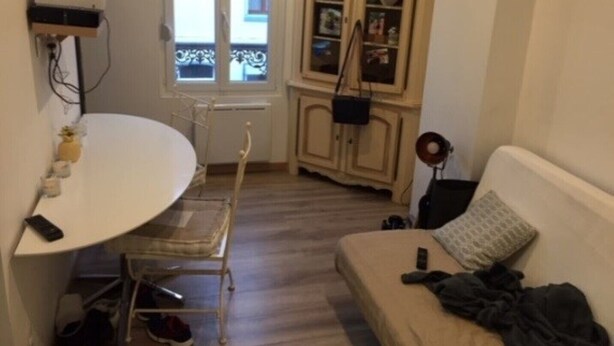 A small room with a white table, a white chair, a beige sofa, a cabinet with glass doors, and a window with a view of buildings outside. - Quiet and bright apartment near the shops (Berck)