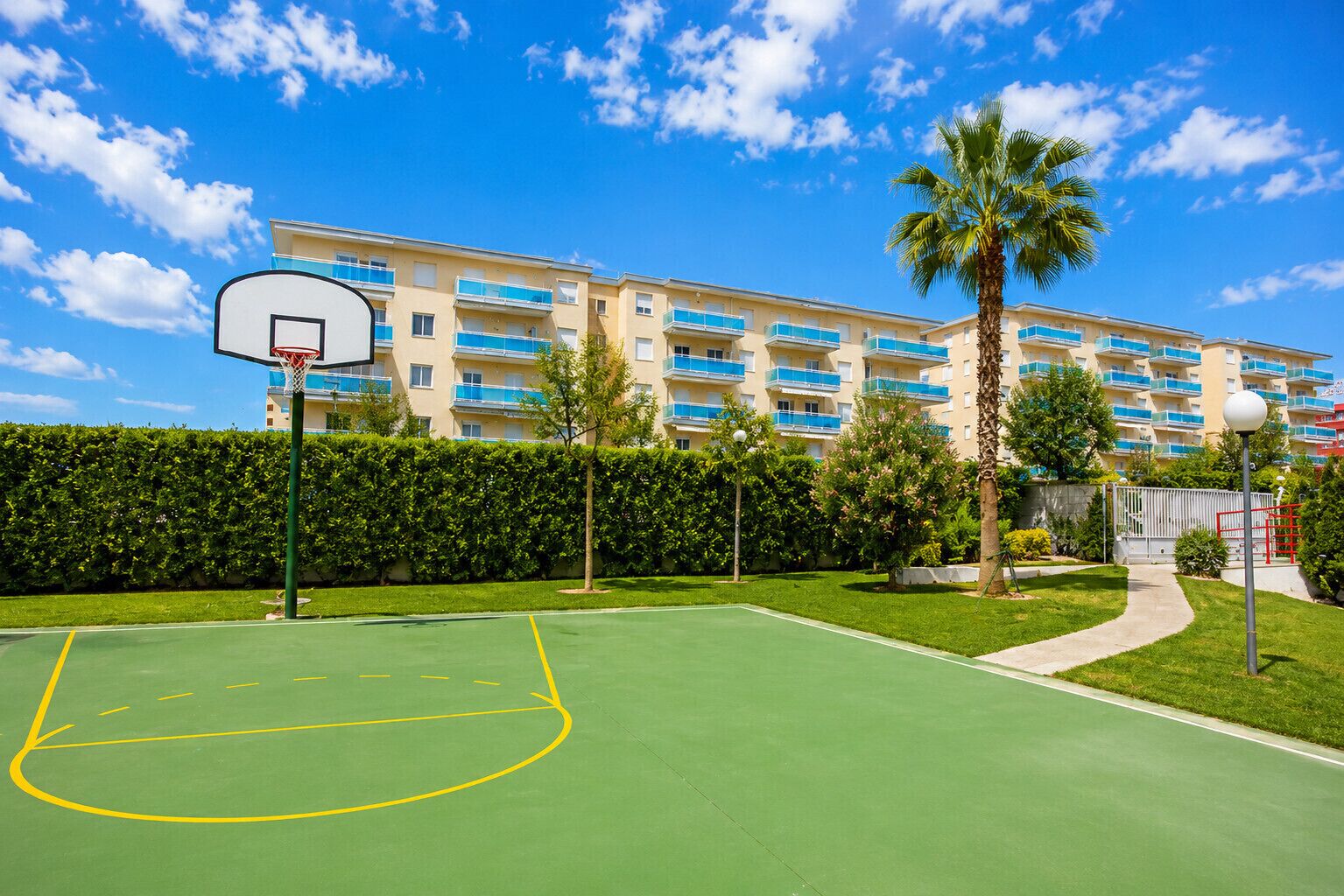 Una cancha de baloncesto con aro, rodeada por un seto y palmeras, con un edificio de varios pisos al fondo.