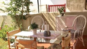 Een terras buiten met rieten stoelen, een glazen tafel, omgeven door groen en een rood hek.