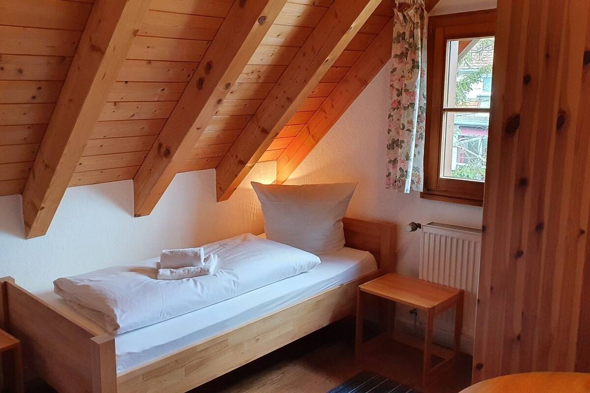 A small, wooden, attic-style bedroom with a single bed, a window with floral curtains, and a radiator.