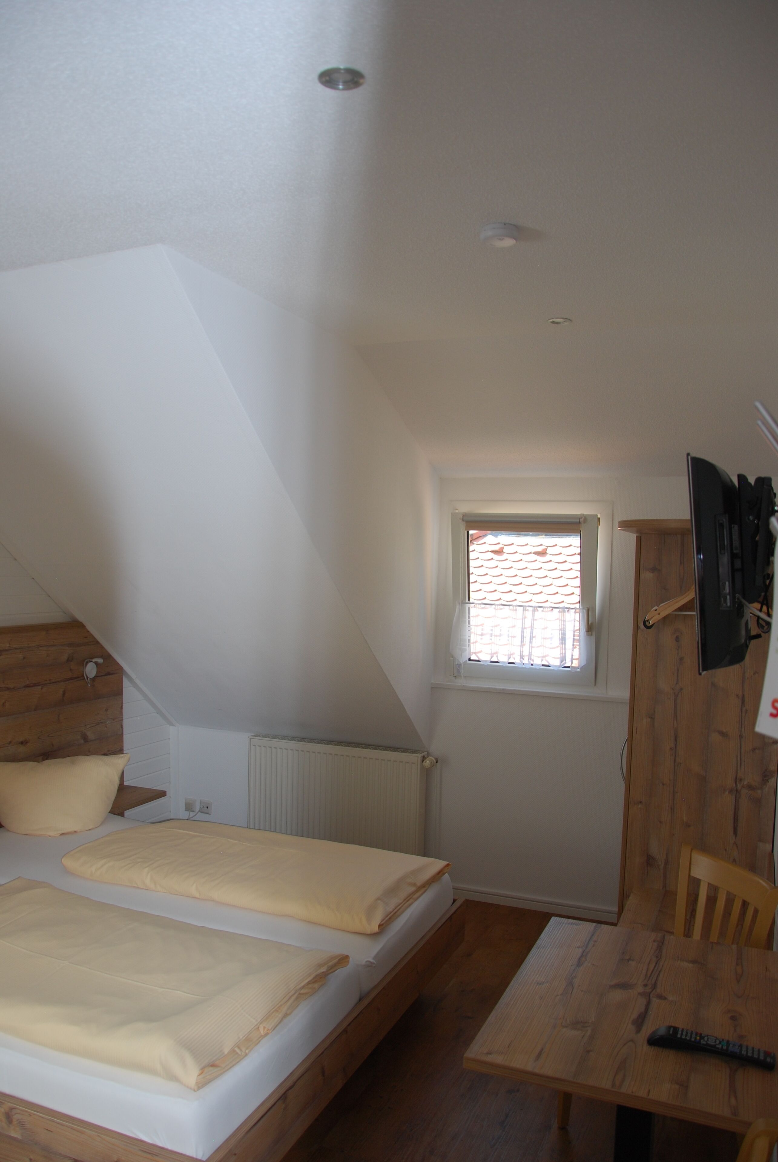 A small, well-lit room with a sloped ceiling, a single window, a bed with two pillows, a wooden table, and a mounted TV.