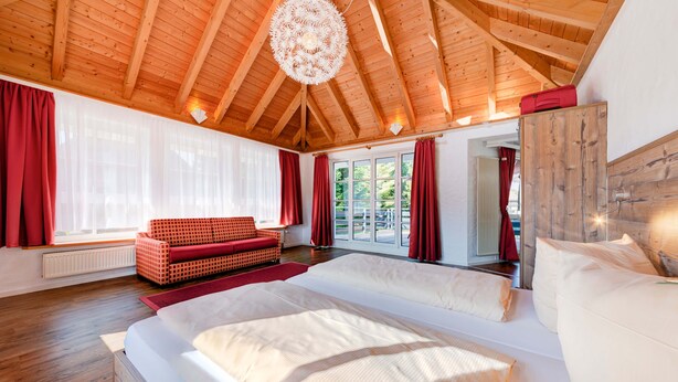 Une chambre spacieuse avec un grand lit, des poutres apparentes en bois au plafond et une vue sur l’extérieur.