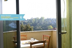 En balkon med et bord og stole, udsigt over et skovområde, samt en dør med gardin i grøn farve.