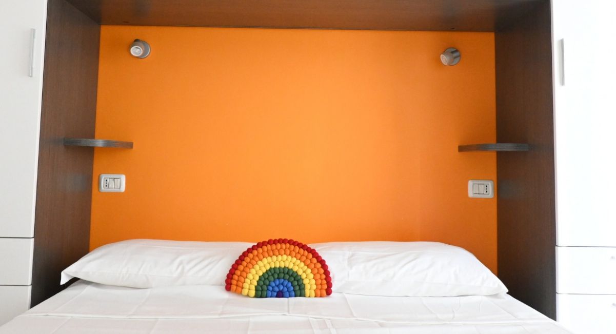 Een bed met een witte matras en kussens, een kleurrijk object op de kussens, en een oranje muur.