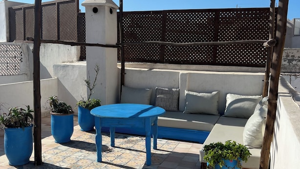 Una terraza en la azotea con una pérgola de madera, un sofá, una mesa azul y plantas en macetas.