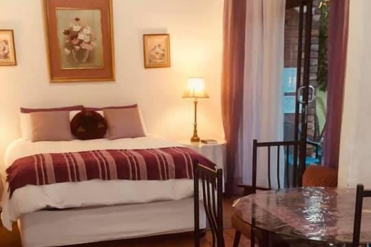 Une chambre à coucher comprenant un lit, une table de chevet, une lampe, une chaise, une table et une fenêtre avec des rideaux.