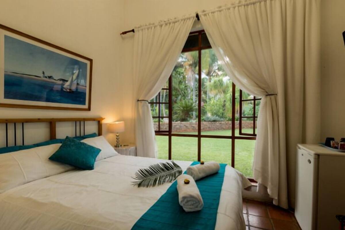 Une chambre à coucher avec un lit, un tableau, des rideaux et une vue sur la verdure à l’extérieur.