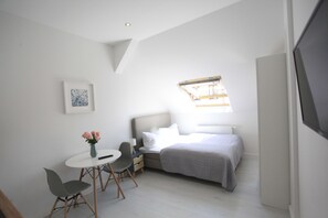 Une chambre à coucher moderne et minimaliste, comprenant un lit, une table de chevet, une chaise et un petit bureau agrémenté d’un vase de fleurs.