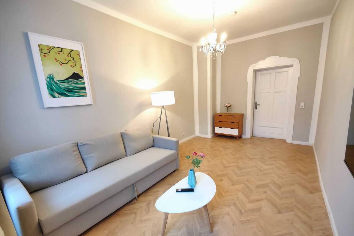 Una sala de estar con un sofá, una pequeña mesa con un florero, una lámpara y una puerta al fondo.