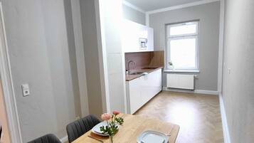 A dining area with a wooden table set for four, a vase with a single flower, and black chairs. A kitchen is visible in the background with white cabinets and a window.